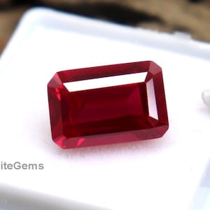 May include: A rectangular, faceted ruby gemstone, presented in a white plastic case. The ruby is a deep red colour and has a polished surface. The case is open, revealing the gem against a white background. The text "EliteGems" is printed on the case.