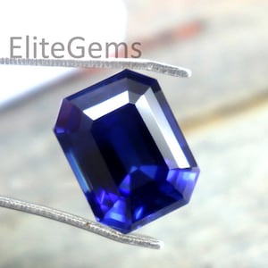 Puede incluir: Una gema azul oscuro, talla esmeralda, es sostenida por unas pinzas plateadas. La gema rectangular tiene esquinas cortadas y facetas que reflejan la luz. El fondo es borroso, de tono neutro.