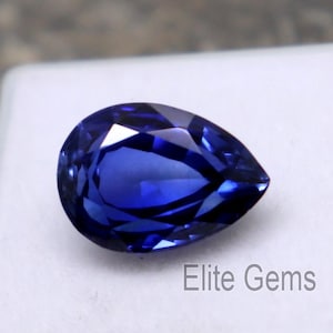 Puede incluir: Una gema azul oscuro facetada en forma de pera. La gema está colocada sobre un fondo blanco, con el texto "Elite Gems" visible. Las facetas de la piedra reflejan la luz, mostrando su claridad y color.