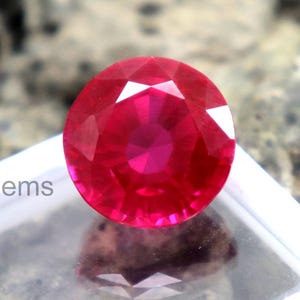 Puede incluir: Una gema roja brillante, probablemente un rubí, de forma redonda y facetada, se muestra en una caja de plástico blanco. La gema tiene una alta claridad y brillo, reflejando la luz. Se lee "Elite Gems" en la caja.