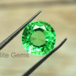 Op de afbeelding: Een ronde, gefacetteerde, heldergroene edelsteen, vastgehouden door een pincet. De steen heeft een briljante slijpvorm, die het licht reflecteert. De tekst "Elite Gems" is zichtbaar. De achtergrond is wazig en neutraal.