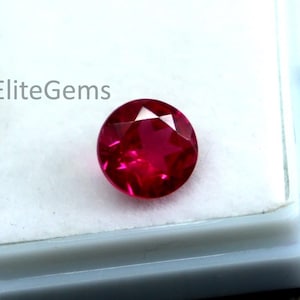 Könnte beinhalten: Ein facettierter, runder, tiefroter Edelstein, wahrscheinlich ein Rubin, liegt in einem weißen Kunststoffetui. Der Edelstein ist der Blickfang und zeigt seinen Brillantschliff und seine satte Farbe. Das Etui befindet sich auf einer dunklen, strukturierten Holzoberfläche.