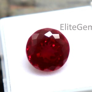 May include: A faceted, round, deep red gemstone sits in a white plastic case. The gem has a brilliant cut, reflecting light. The case is open, revealing the gem against a natural wood background. The text "EliteGems" is visible on the case.