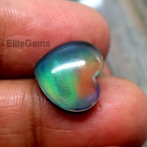 Puede incluir: Una piedra preciosa en forma de corazón con una vibrante exhibición de colores, incluyendo verde, azul, naranja y púrpura. La gema se sostiene en una mano, mostrando sus cualidades iridiscentes y su superficie pulida. La gema es de EliteGems.