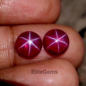 Könnte beinhalten: Zwei runde, tiefrote Edelsteine mit einem sechsstrahligen Sternmuster. Die Edelsteine werden in einer Hand gehalten, wodurch ihre polierten Oberflächen und der Sterneffekt zur Geltung kommen. Der Text "EliteGems" ist sichtbar.