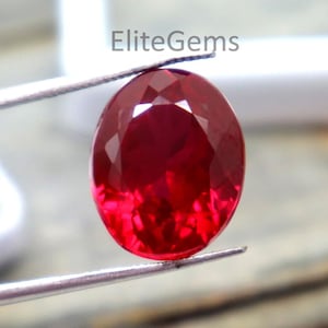 May include: A faceted, oval-shaped, deep red gemstone held by tweezers. The gem has a brilliant cut, reflecting light. The background includes a white container and the text "EliteGems".