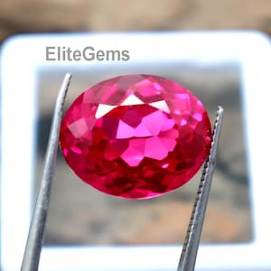Könnte beinhalten: Ein leuchtend rosa, oval geschliffener Edelstein wird von einer silbernen Pinzette gehalten. Der facettierte Stein reflektiert das Licht vor einem weißen Hintergrund. Der Text "EliteGems" ist sichtbar. Es handelt sich wahrscheinlich um einen Rubin oder rosa Saphir.