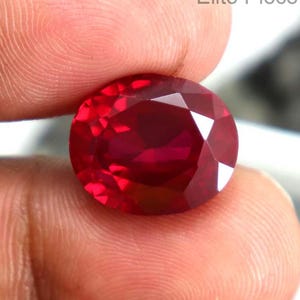 May include: A faceted, oval-shaped ruby gemstone. The ruby is a deep, vibrant red color and is held between fingers. The gem is displayed against a white background. The text "Elite Piece" is visible.
