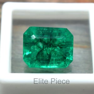 May include: A vibrant green emerald gemstone, rectangular cut, displayed in a white plastic case. The gemstone has visible internal features and is set against a neutral background. The text "Elite Piece" is printed on the case.
