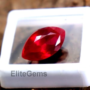 Peut inclure: Une pierre précieuse rouge vif, taillée en forme ovale, est présentée dans un boîtier en plastique blanc. La gemme a un éclat élevé et est posée sur un fond en bois naturel. Le texte "EliteGems" est imprimé sur le boîtier.