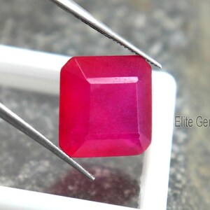 May include: A vibrant, square-cut, ruby-red gemstone is held by tweezers above a white display case. The gem has a polished surface and is set against a textured gray background. The text "Elite Gems" is visible.
