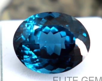 Puede incluir: Una gema facetada, de forma ovalada y color verde azulado. La gema está colocada sobre una superficie blanca, con el texto "ELITE GEMS" visible. El fondo es una formación rocosa borrosa de tonos neutros. Las facetas de la gema reflejan la luz.