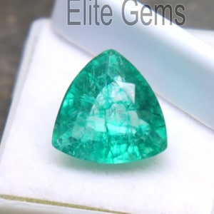 Puede incluir: Una piedra preciosa de esmeralda triangular y facetada, exhibida en un estuche de plástico blanco. La gema es de color verde vibrante y tiene una superficie pulida. El texto "Elite Gems" es visible en el fondo.