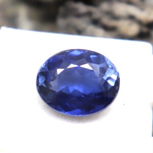 Puede incluir: Una gema azul oscuro, facetada y de forma ovalada, descansa sobre una superficie blanca. La gema tiene un corte brillante, que refleja la luz. La gema se encuentra sobre un fondo neutro, resaltando su color y claridad. Este es un componente de joyería.