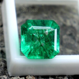 Puede incluir: Una vibrante gema verde talla esmeralda se muestra en una caja cuadrada blanca. La gema tiene una apariencia brillante y clara con inclusiones internas. El texto "Elite Gems" es visible en la caja. El fondo es una superficie oscura y texturizada.