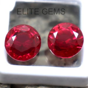 Puede incluir: Dos gemas redondas, facetadas, de color rojo intenso se muestran en una caja de plástico blanco. Las gemas tienen un corte brillante, que refleja la luz. El texto "ELITE GEMS" es visible en la caja. El fondo es una superficie texturizada gris.