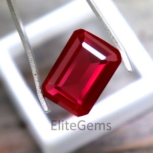 May include: A rectangular-cut ruby gemstone, held by tweezers, is shown in a white square. The ruby's deep red colour is highlighted by its facets, reflecting light. The text "EliteGems" is visible.