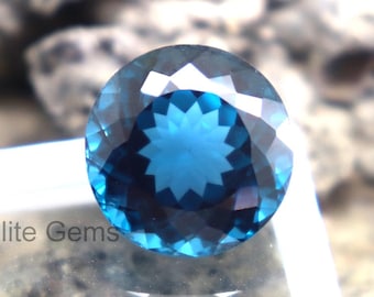 Puede incluir: Una gema facetada, redonda, de color verde azulado oscuro. La gema está colocada en una caja de plástico transparente, con el texto "Elite Gems" visible. El fondo es una superficie borrosa, texturizada, de color marrón claro y gris.