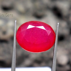 May include: A faceted, oval-shaped, bright red gemstone is held by silver tweezers. The gem has a polished, glossy surface and a white, geometric pattern on top. The text "Elite Gems" is visible in the background.