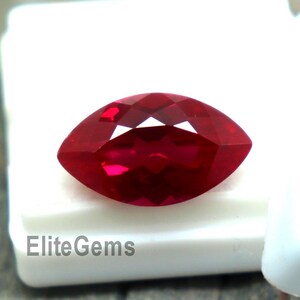May include: A faceted, marquise-cut ruby gemstone, displayed in a white box. The ruby is a deep, vibrant red color, with multiple facets catching the light. The text "Elite Gems" is printed on the box.