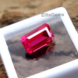 Peut inclure: Une pierre précieuse rouge vif, rectangulaire et à facettes est présentée dans un boîtier en plastique blanc. La pierre est posée sur un fond sombre. Le texte "EliteGems" est visible sur le boîtier.