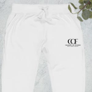 以下が含まれることがあります： 白いスウェットパンツ、ドローストリングウエストとポケット付き。パンツには「COF CHANNEL OF FLYNES！MUSIC. GAMING. CREATIVE.」と書かれた黒いロゴが付いています。