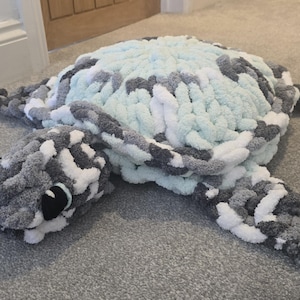 Könnte beinhalten: Ein großes, handgefertigtes kissenförmiges Schildkröte in Grau-, Weiß- und Hellblautönen. Das Plüschkissen hat ein strukturiertes, grobes Strickdesign mit detailliertem Kopf und Flossenform. Die Augen der Schildkröte sind schwarz und weiß.