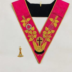 Puede incluir: Un collar y un colgante ceremoniales. El collar es de color rosa vibrante con diseños florales bordados en oro y verde y un reverso negro. También hay un pequeño colgante dorado con detalles en rojo y blanco.