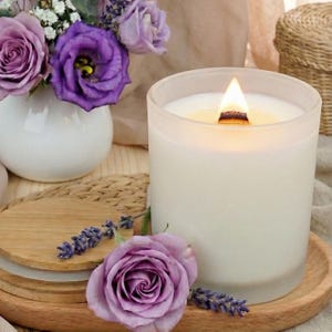 Peut inclure: Une bougie blanche allumée dans un récipient en verre dépoli avec une mèche en bois. La bougie est posée sur un plateau en bois avec une rose violette et des brins de lavande. Un petit vase blanc avec des fleurs violettes est en arrière-plan.