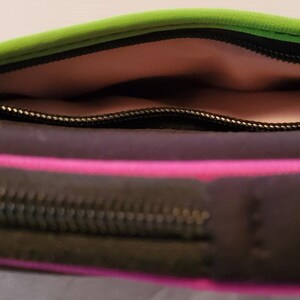 Pu&ograve; includere: Una pochette con cerniera nera, verde e rosa con fodera rosa. La pochette &egrave; aperta e la cerniera &egrave; visibile.