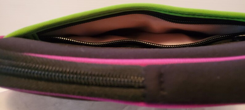 Pu&ograve; includere: Una pochette con cerniera nera, verde e rosa con fodera rosa. La pochette &egrave; aperta e la cerniera &egrave; visibile.
