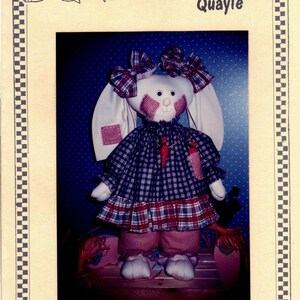 May include: A handmade patchwork bunny doll with a red and blue plaid dress, white ears, and a red bow. The doll is standing on a wooden surface. The image is titled "The Country Quayle" and "Patches".