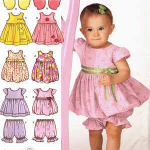May include: A sewing pattern for a baby girl's dress and bloomers. The pattern includes five different styles, including a yellow dress with buttons, a pink dress with a flower, a floral print dress, a pink gingham dress with a green sash, and a purple dress with a flower. The pattern is from Simplicity and is size A XXS, XS, S, M, L. The pattern number is 3854.