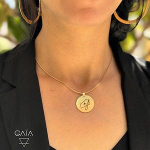 Op de afbeelding: Een goudkleurige ketting met een ronde hanger met een geometrisch ontwerp en een ster. De ketting wordt gedragen met grote gouden oorringen en een zwarte blazer. Het merklogo "GAIA" is onderaan zichtbaar.
