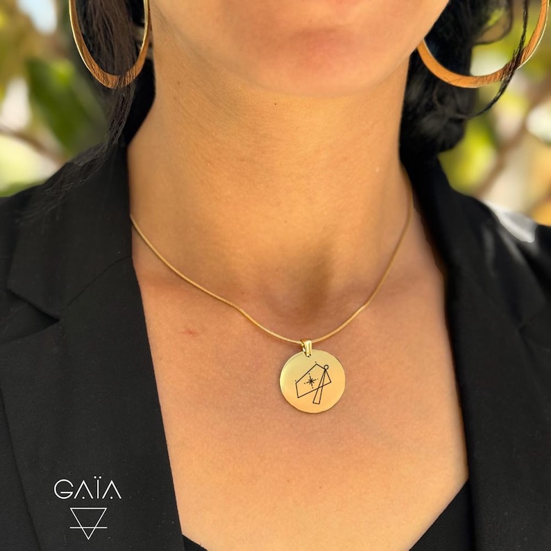 Op de afbeelding: Een goudkleurige ketting met een ronde hanger met een geometrisch ontwerp en een ster. De ketting wordt gedragen met grote gouden oorringen en een zwarte blazer. Het merklogo "GAIA" is onderaan zichtbaar.