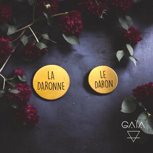 Peut inclure: Deux dessous de verre ronds dorés avec les inscriptions noires "LA DARONNE" et "LE DARON". Posés sur une surface sombre, ils sont entourés de fleurs bordeaux et de feuilles vertes. Le logo "GAIA" est en bas à droite.
