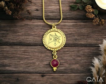 Op de afbeelding: Een goudkleurige hangerketting met een cirkelvormige bedel met een geëtst geometrisch ontwerp en een kleine, ronde, rode edelsteen. De ketting wordt getoond op een houten oppervlak met gedroogde planten en een brandende kaars op de achtergrond.