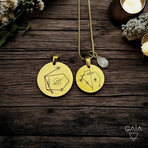 Op de afbeelding: Twee goudkleurige, ronde hangerkettingen met geometrische ontwerpen. E&eacute;n hanger heeft een pijl en een zonachtig symbool, de andere een ster- en lijnenontwerp. E&eacute;n ketting heeft een druppelvormige, lichtgekleurde steen. De hangers hangen aan dunne gouden kettingen.