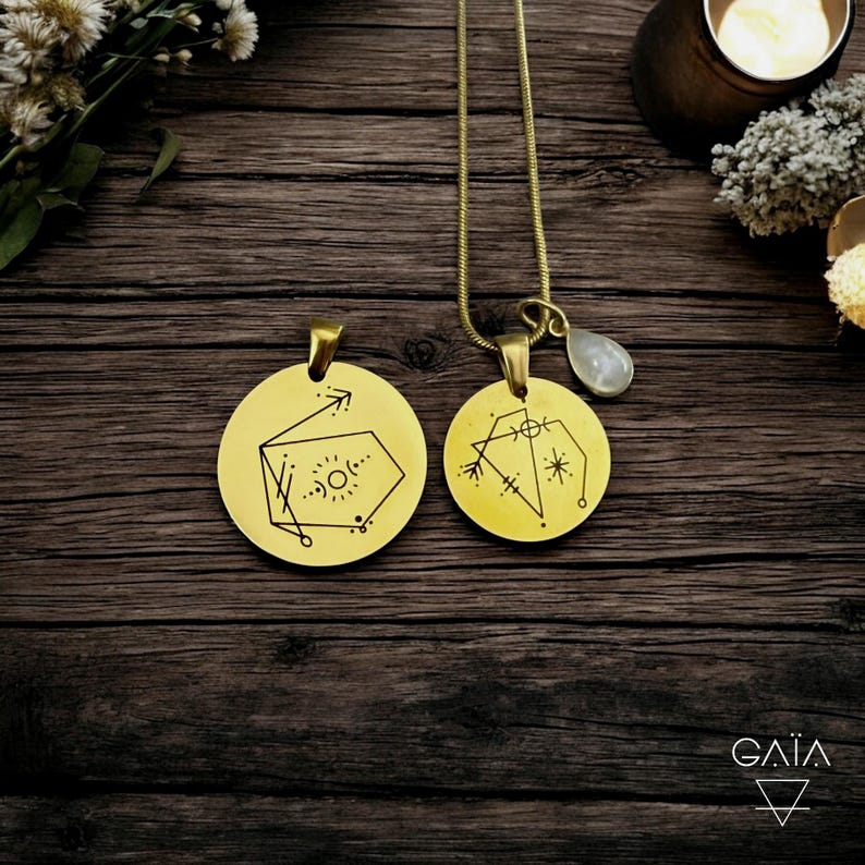 Op de afbeelding: Twee goudkleurige, ronde hangerkettingen met geometrische ontwerpen. E&eacute;n hanger heeft een pijl en een zonachtig symbool, de andere een ster- en lijnenontwerp. E&eacute;n ketting heeft een druppelvormige, lichtgekleurde steen. De hangers hangen aan dunne gouden kettingen.