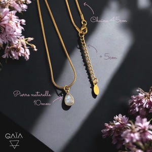 Op de afbeelding: Een goudkleurige ketting met een druppelvormige natuurstenen hanger. De ketting is 45 cm lang, met een verlenging van 5 cm. De steen meet 10 mm. De ketting wordt gepresenteerd op een donkere ondergrond met roze bloemen.