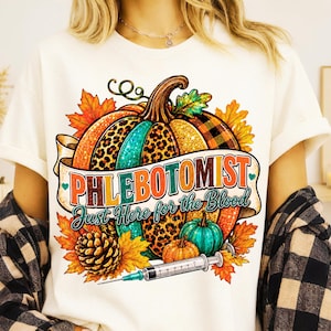 Puede incluir: Una camiseta color crema con un gráfico de una calabaza grande con secciones de estampado de leopardo y purpurina, rodeada de hojas de otoño y una piña. El texto dice PHLEBOTOMIST Just Here for the Blood, con una jeringa médica debajo.