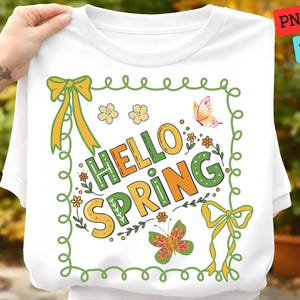 Può includere: T-shirt bianca con grafica colorata "Hello Spring". Il design presenta farfalle, fiori e fiocchi nei toni del giallo, verde e arancione. Il testo è delineato in verde e arancione, con un bordo verde.