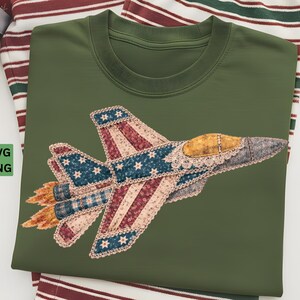 May include: Olive green crew neck t-shirt featuring an appliqued jet plane design. The jet has a patriotic color scheme with red, white, and blue fabric, and a yellow cockpit. The design includes stars and stripes.