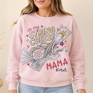 Puede incluir: Una sudadera de cuello redondo rosa claro con un gráfico de queso en tiras saliendo de su envoltorio. El texto dice in my STRING CHEESE MAMA era, junto con la frase Overwhelmed, touched out, but exactly who I'm meant to be.