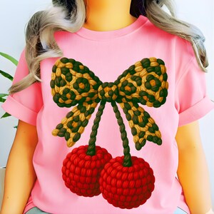 Könnte beinhalten: Ein hellrosa T-Shirt mit einem großen Grafikdruck von zwei roten, texturierten, kugelförmigen Früchten, die an Stielen hängen, welche an einer zentralen Schleife befestigt sind. Die Schleife hat ein beiges und dunkelbraunes Leopardenmuster in Seiloptik.