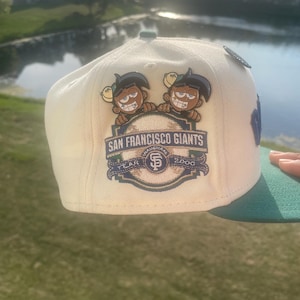 May include: Cream-colored baseball cap with a green brim. The cap features a San Francisco Giants logo with cartoon characters and the text "SAN FRANCISCO GIANTS" and "YEAR 2000".