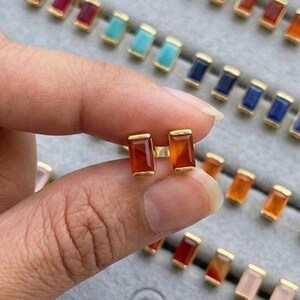May include: A pair of gold-colored stud earrings with rectangular, reddish-orange gemstones. The earrings are held in a hand, with a tray of similar earrings in various colors in the background. The jewelry is from the brand "JEWENLERY JEWELRY".