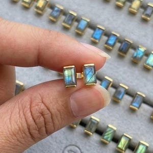 May include: Gold-tone rectangular stud earrings with iridescent blue-green stones. The earrings are held in a hand, with several pairs of similar earrings arranged in a gray jewelry tray. The tray has the text "JEWENLERY JEWELRY" printed on it.