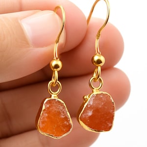 Könnte beinhalten: Ein Paar goldfarbene Ohrringe mit orangefarbenen, unregelmäßig geformten Steinen. Die Steine sind in goldfarbenem Metall gefasst und hängen an goldfarbenen Ohrhaken. Die Ohrringe werden von einer Hand vor weißem Hintergrund gehalten.