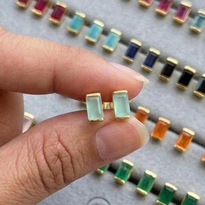 May include: A pair of light green rectangular stud earrings with gold-colored settings. The earrings are held in a hand, with a tray of similar earrings in various colors in the background. The jewelry is from the brand "JEWENLERY JEWELRY."